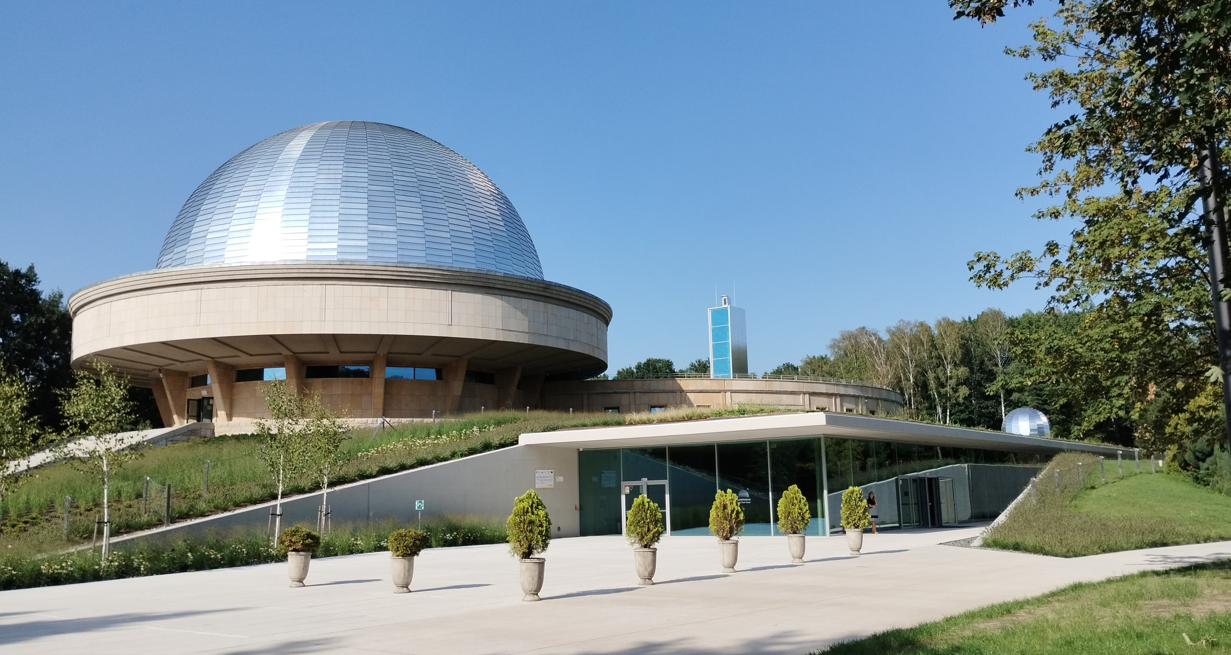 Planetarium Śląskie w Chorzowie: kopuła sali projekcyjnej przypominająca połowę planety Saturn z pierścieniami, wejście do nowowybudowanej części z kawiarnią i wystawami tematycznymi, donice z tujami na placu przed wejściem, w tle wieża widokowa oraz obserwatorium astronomiczne im. Jana Palta PTMA.